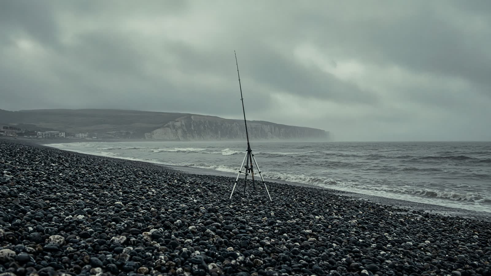 A UK shingle beach under an overcast sky, with a beachcaster in a tripod at the tide line.